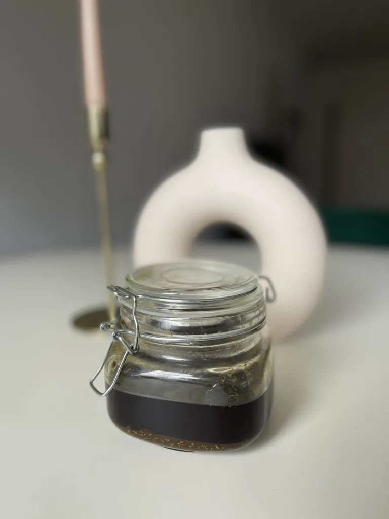 A glass jar of hair growth oil is sitting on a white table. The oil is greenish in color.