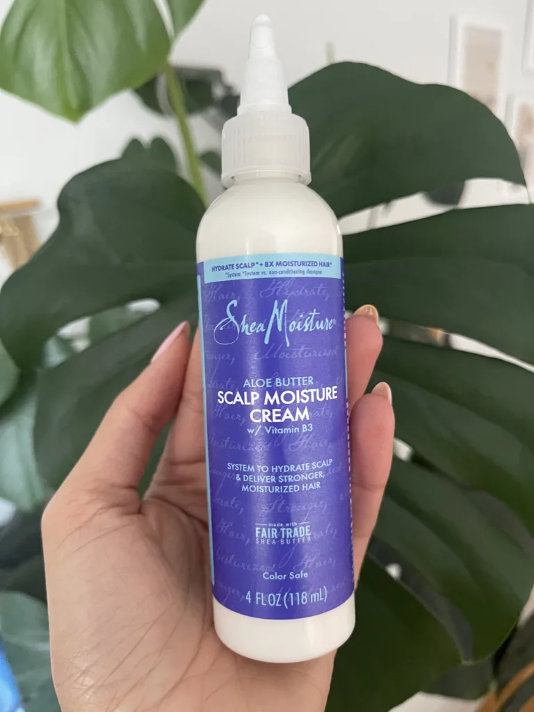 Hand holding a bottle of SheaMoisture's Aloe Butter Scalp Moisture Cream. The slim bottle has a purple label with light blue writing which reads: SheaMoisture Aloe Butter Scalp Moisture Cream w/ Vitamin B3. System to Hydrate Scalp & Deliver Stronger, Moisturized Hair.