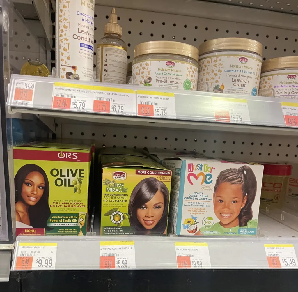Three boxes of hair relaxers – ORS Olive Oil Relaxer, African Pride Olive Oil Miracle Relaxer, and Just For Me No-Lye Relaxer – displayed on the bottom shelf of the beauty aisle in a convenience store.