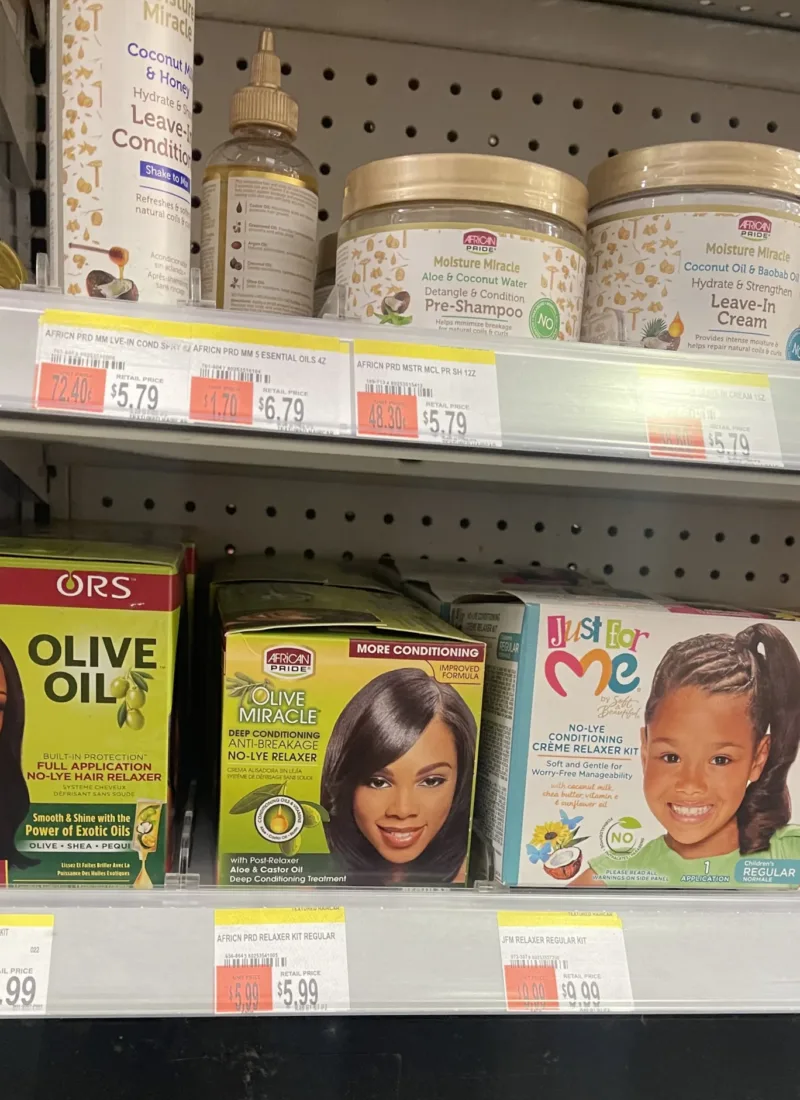 Three boxes of hair relaxers – ORS Olive Oil Relaxer, African Pride Olive Oil Miracle Relaxer, and Just For Me No-Lye Relaxer – displayed on the bottom shelf of the beauty aisle in a convenience store.