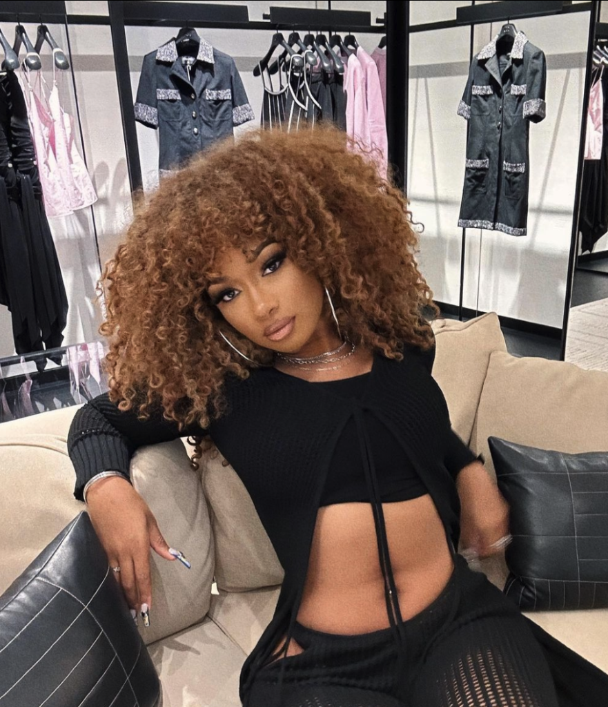 Rapper Megan Thee Stallion, with curly honey blonde hair, wearing a black outfit sitting on a tan couch.