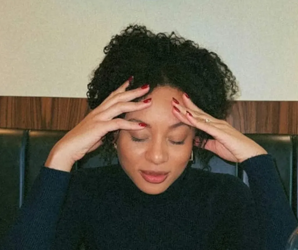 A tan woman with curly hair touching the sides of her head as if she has a headache.