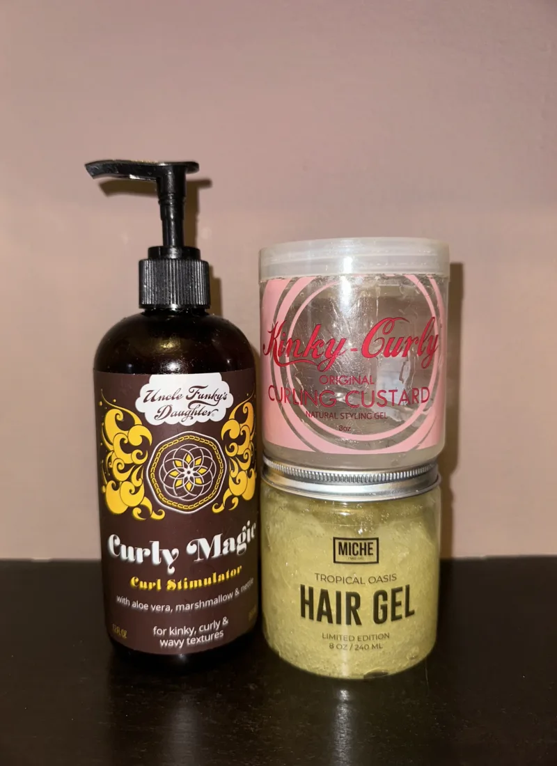 A close-up photo of three hair care products: Uncle Funky's Daughter Curly Magic Curling Custard, Miche Tropical Oasis Natural Styling Gel, and Curt Hair Gel. The products are arranged on a wooden table.
