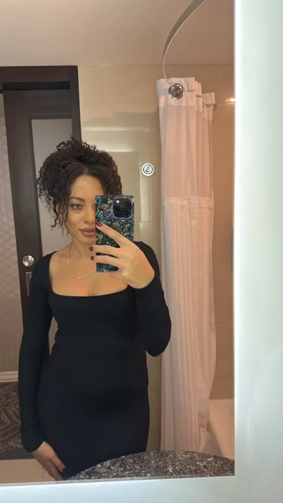 A woman with her curly hair up, wearing a black dress, taking a selfie in a mirror.