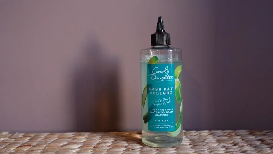 A white and teal bottle of Carol's Daughter Wash Day Delight Shampoo with a blue flip-top lid, sitting on a woven wicker basket. The text on the bottle says "Carol's Daughter", "Wash Day Delight", "Water-to-Foam Shampoo", "with Aloe", "Hair So Clean, Surprisingly Soft", and "Love at First Wash".
