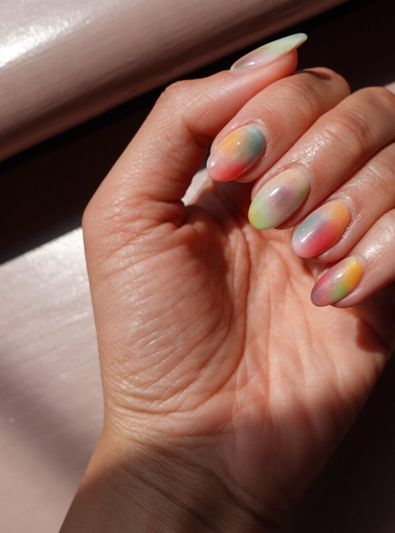 A close-up photo of a woman’s hand with colorful fingernails in various shades.