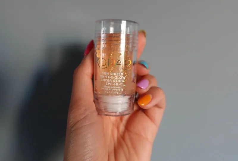 A woman with long, colorful nails holds a clear plastic tube containing an orange-yellow, sparkly liquid. The text on the tube reads "KOPARI SUN SHIELD ON-THE-GLOW SHEER STICK SPF 40"
