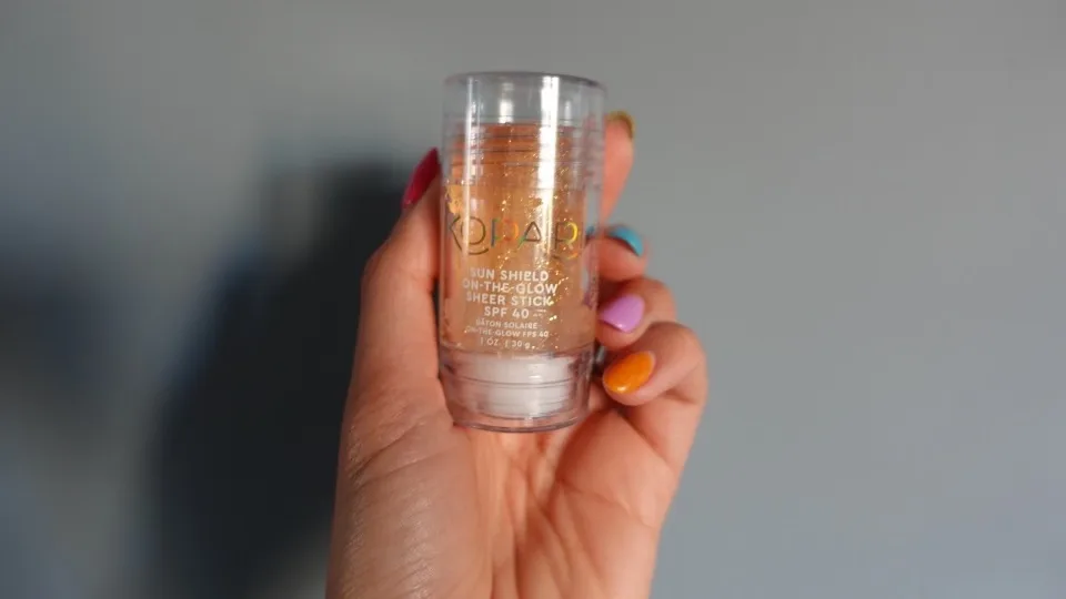 A woman with long, colorful nails holds a clear plastic tube containing an orange-yellow, sparkly liquid. The text on the tube reads "KOPARI SUN SHIELD ON-THE-GLOW SHEER STICK SPF 40"