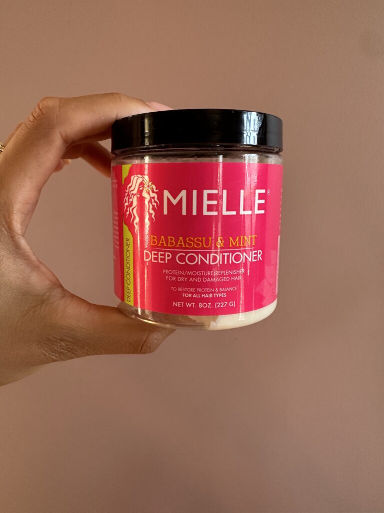 A hand holding a jar of Mielle Organics Babassu Oil & Mint Deep Conditioner against a peach-colored background. The label is pink with black text highlighting the product's benefits for dry and damaged hair.