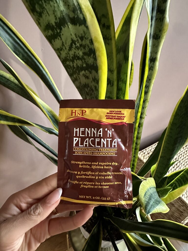 A hand holds a small packet of HNP Henna 'N' Placenta Conditioning Treatment against a backdrop of a lush green snake plant. The packet is brown with orange and yellow accents, and the text describes the product's ability to strengthen and repair dry, brittle, and lifeless hair.