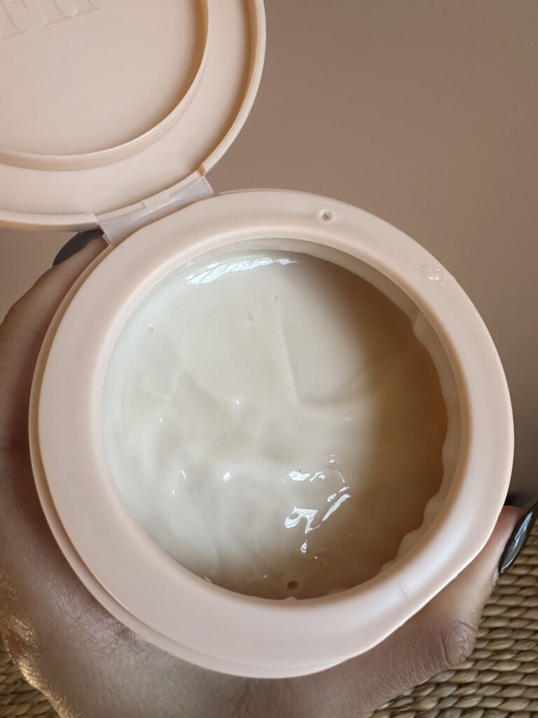 A person's hand holding a pink jar of Fenty Beauty The Homecurl Curl-Defining Cream, with the lid open revealing the creamy texture of the product.